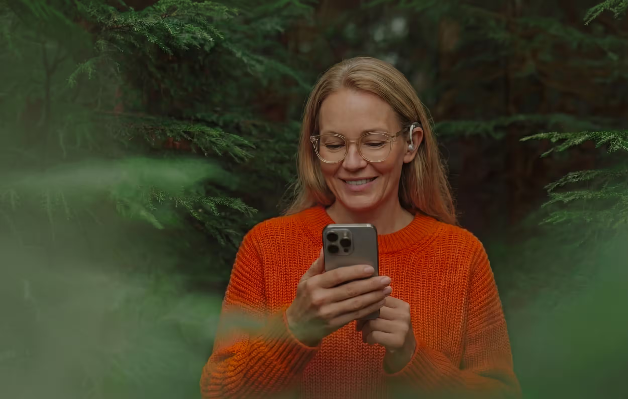 Smiling woman wearing glasses and an orange sweater, holding a smartphone in a forest setting.