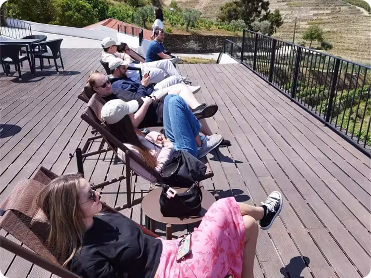 Group of six people lounging on reclining chairs on a wooden deck overlooking a scenic green landscape and terraced hills.