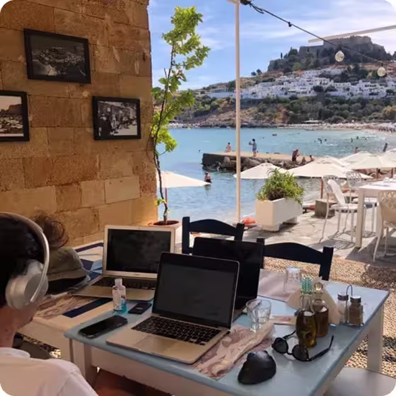 Person wearing headphones working on a laptop at a table outside overlooking a seaside with umbrellas, chairs, and a rocky hill with white buildings.