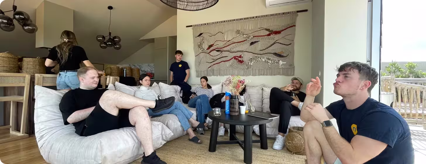 Group of seven young adults relaxing and interacting in a modern living room with white sofas, a black coffee table, and woven baskets.