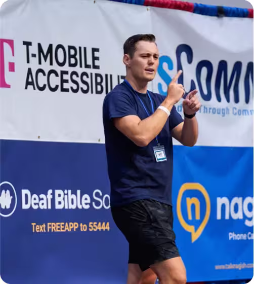 A man in a navy shirt is using sign language in front of banners for T-Mobile Accessibility and Deaf Bible Society.