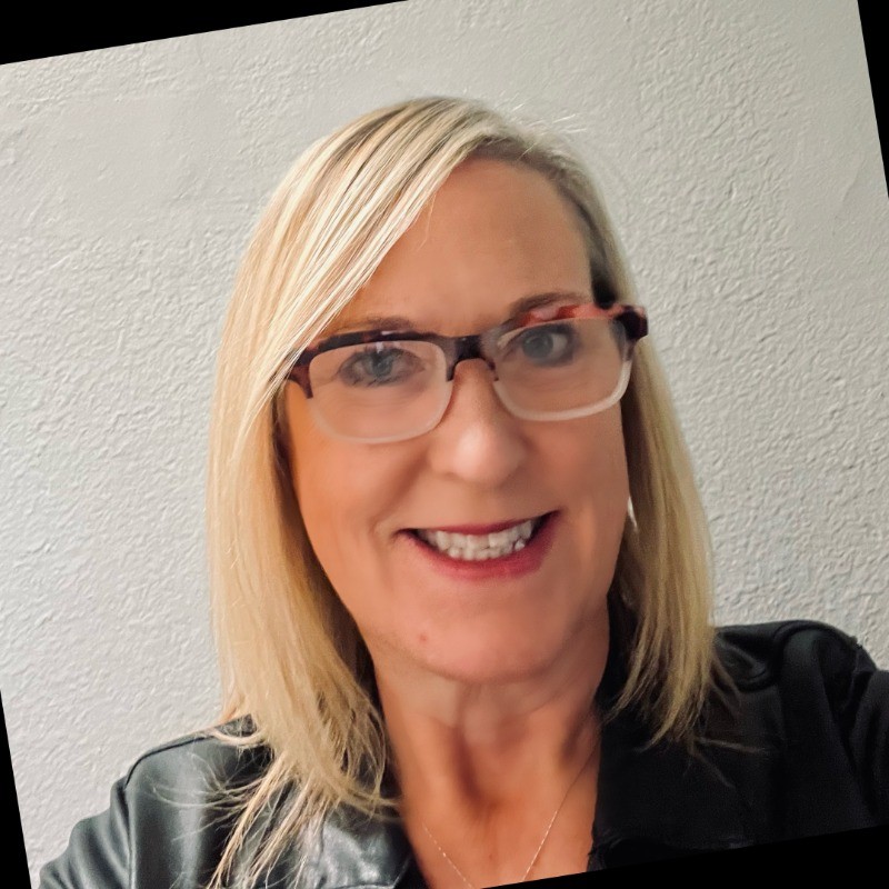 Smiling woman with blonde hair wearing glasses and a black leather jacket against a white textured wall.