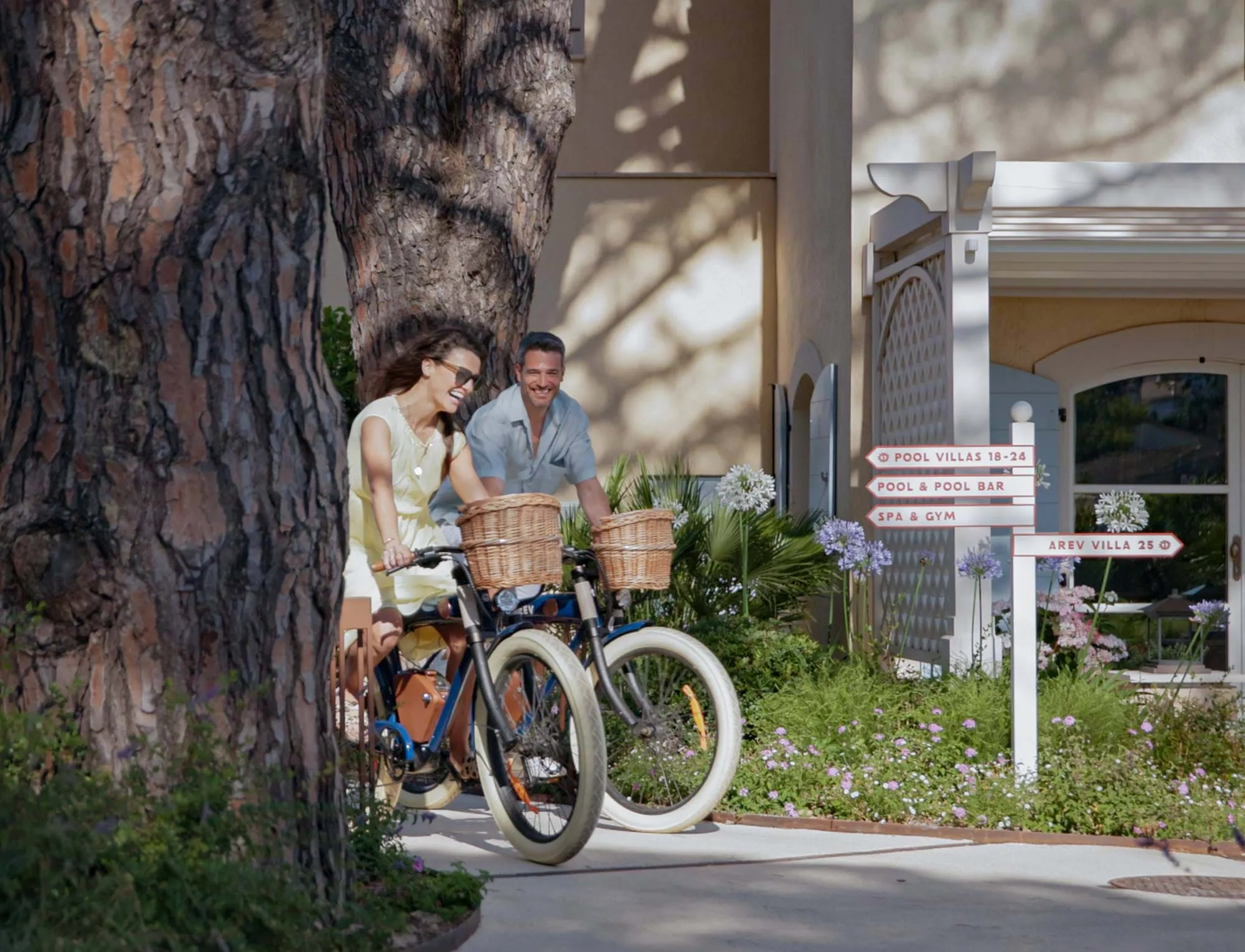 Casal feliz andando de bicicleta com cestas de vime ao longo de um caminho de jardim perto de uma placa indicando moradias na piscina, bar de piscina, spa, ginásio e uma villa.