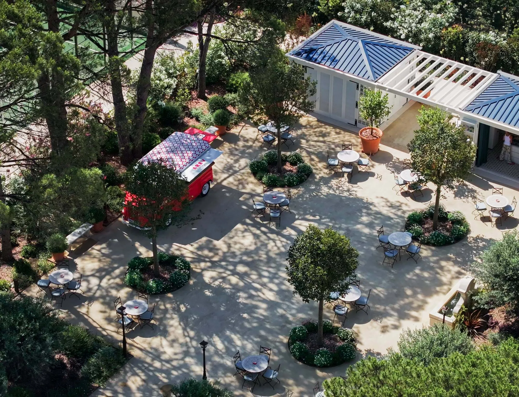 Área de estar ao ar livre do café com mesas redondas, cadeiras, árvores e um carrinho de comida vermelho perto de um edifício branco com telhados azuis.