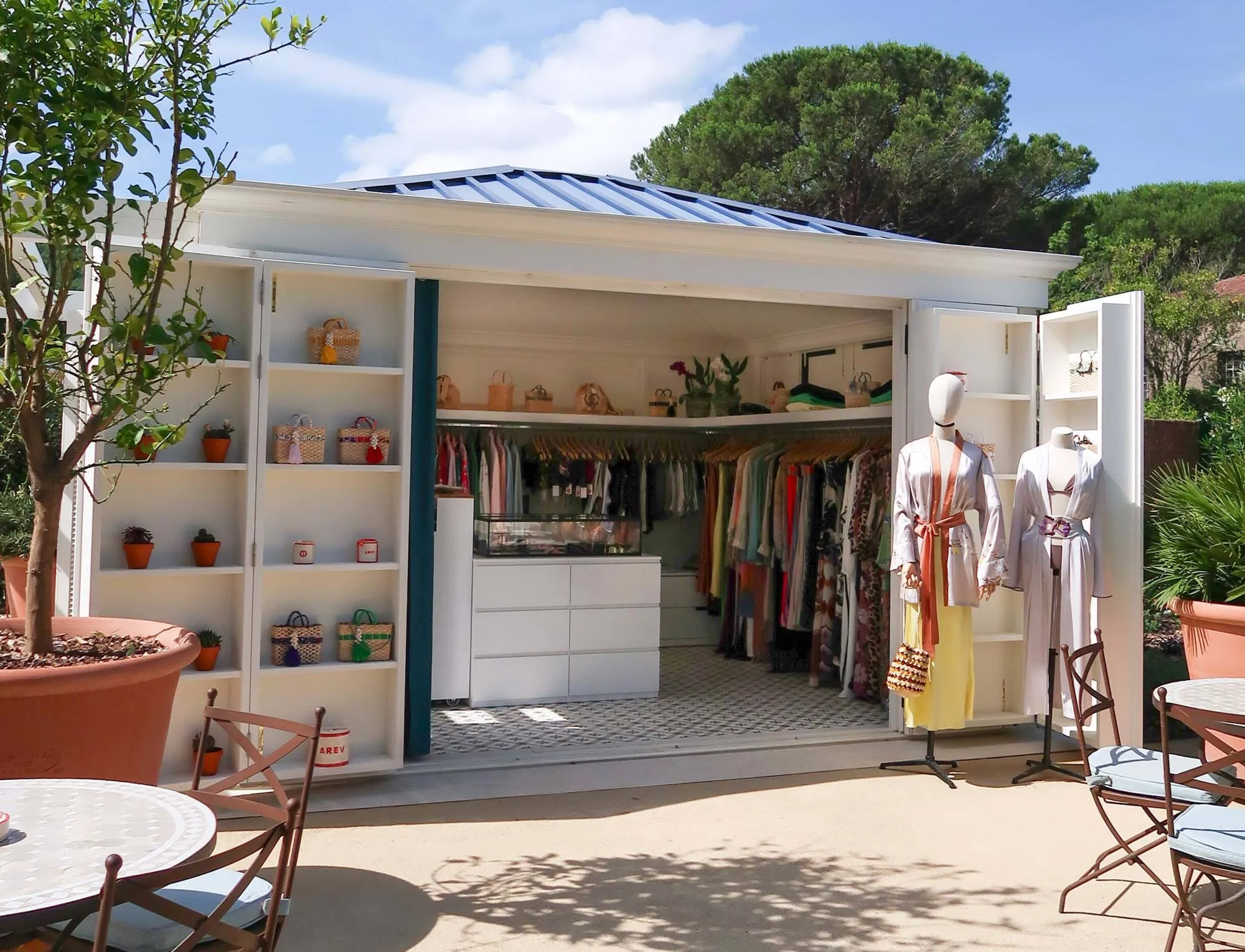 Boutique ao ar livre com armários brancos abertos exibindo cestos, manequins vestidos com roupões e prateleiras de roupas no interior.