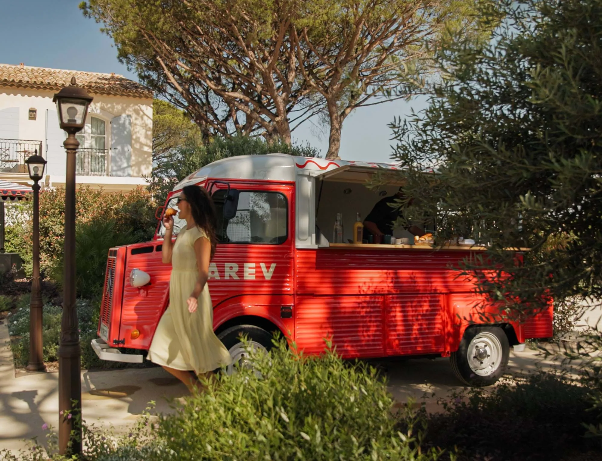 Mulher de vestido amarelo a comer uma pastelaria a passar por um food truck vintage vermelho num jardim ensolarado.