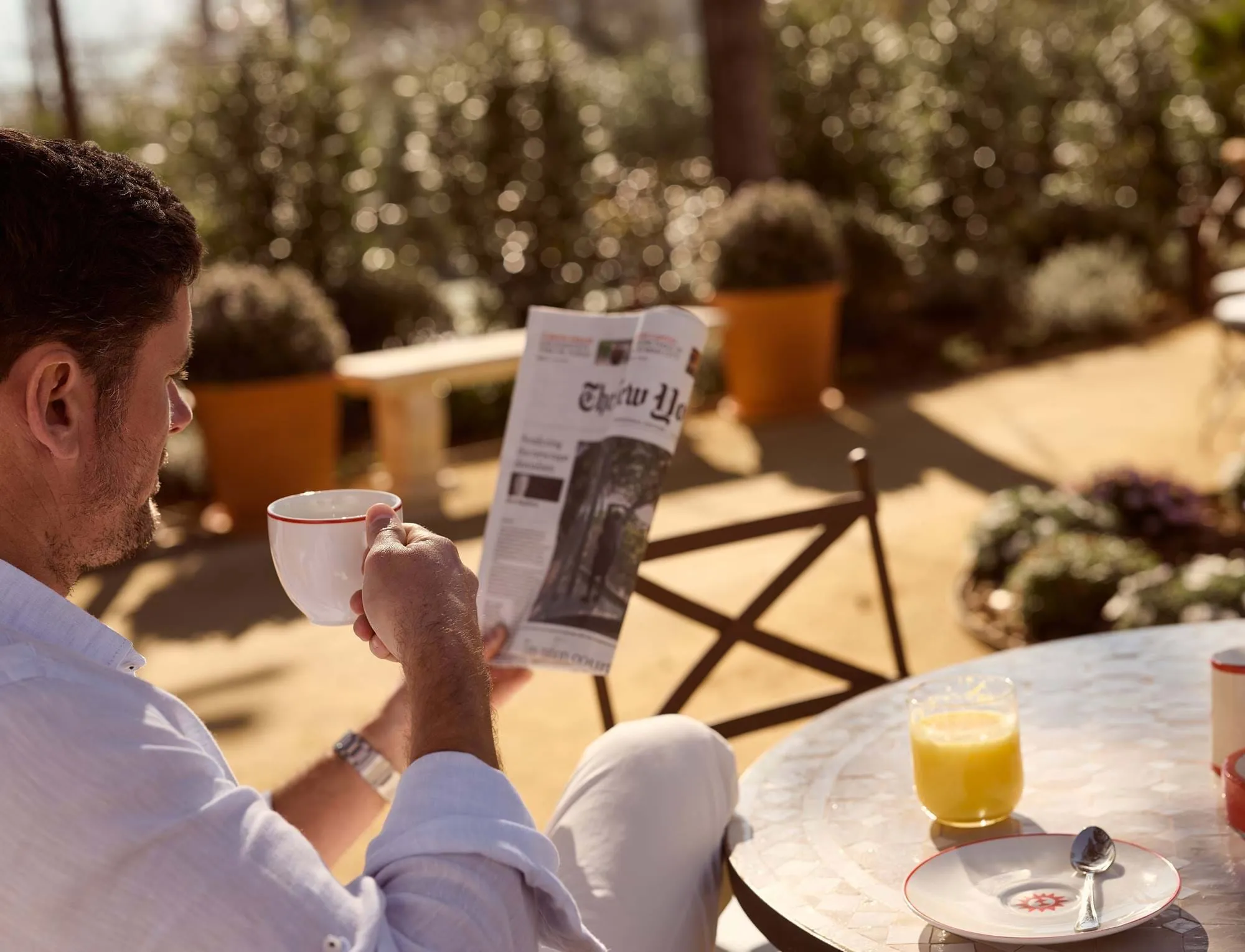 Homem de camisa branca sentado ao ar livre numa mesa redonda, a beber café e a ler um jornal.