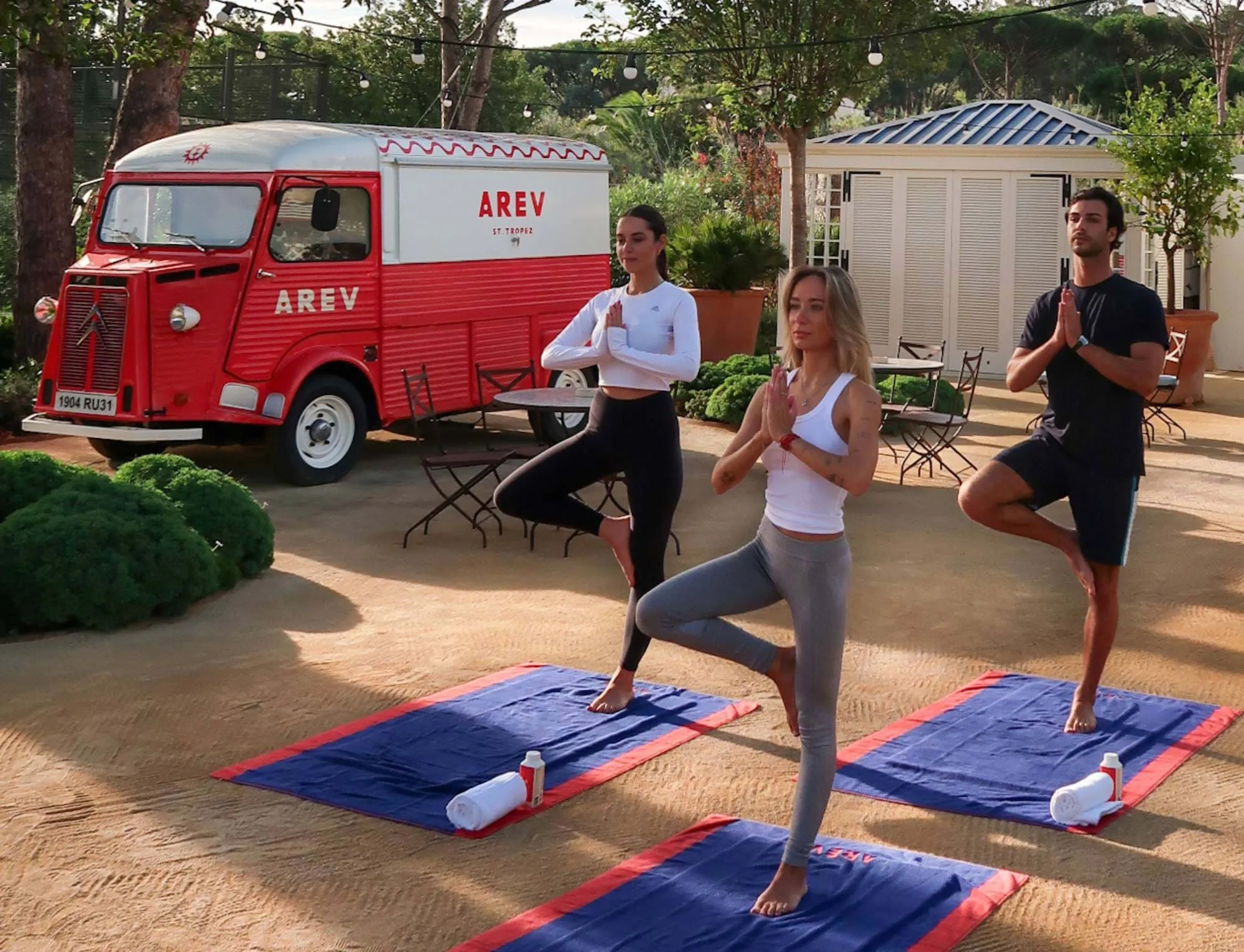 Três pessoas que praticam ioga fazem pose em tapetes azuis ao ar livre perto de uma carrinha AREV vermelha e branca.