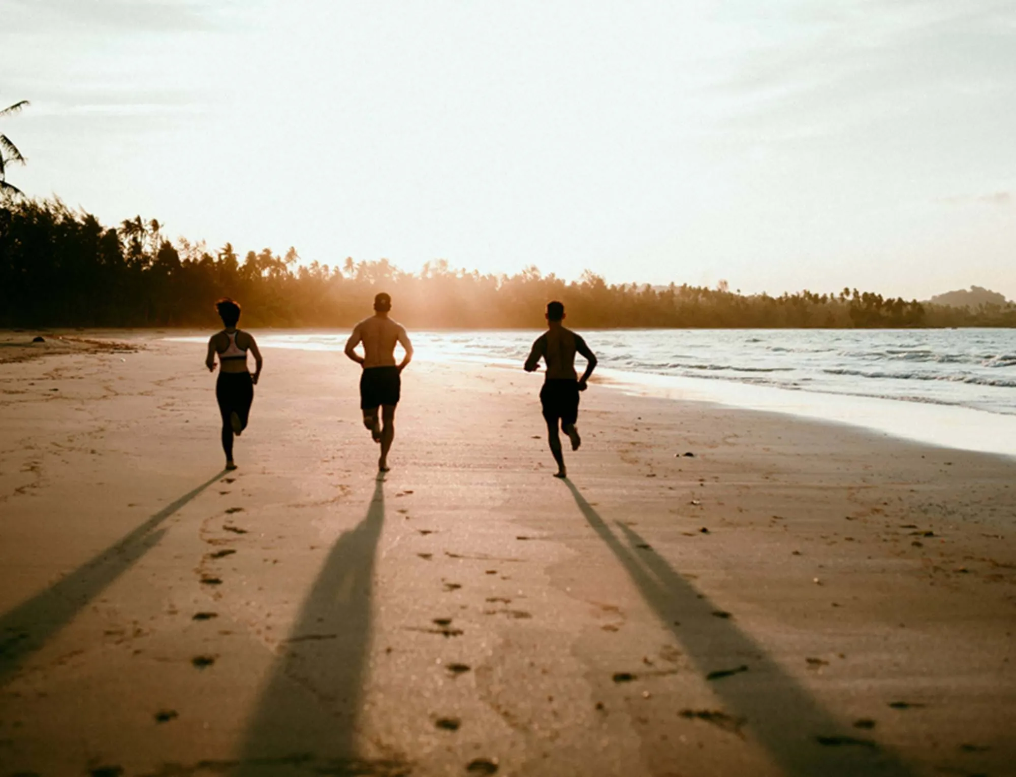 Drei Menschen rennen bei Sonnenuntergang an einem Strand, hinter ihnen erstrecken sich lange Schatten.