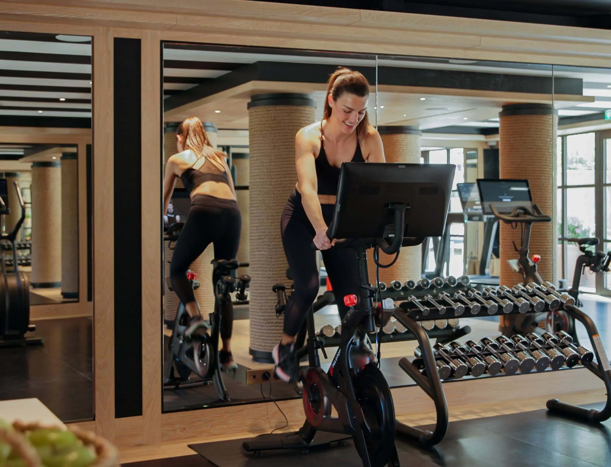 Frau in schwarzer Trainingskleidung trainiert auf einem Heimtrainer in einem modernen Fitnessstudio mit Hanteln und verspiegelten Wänden.