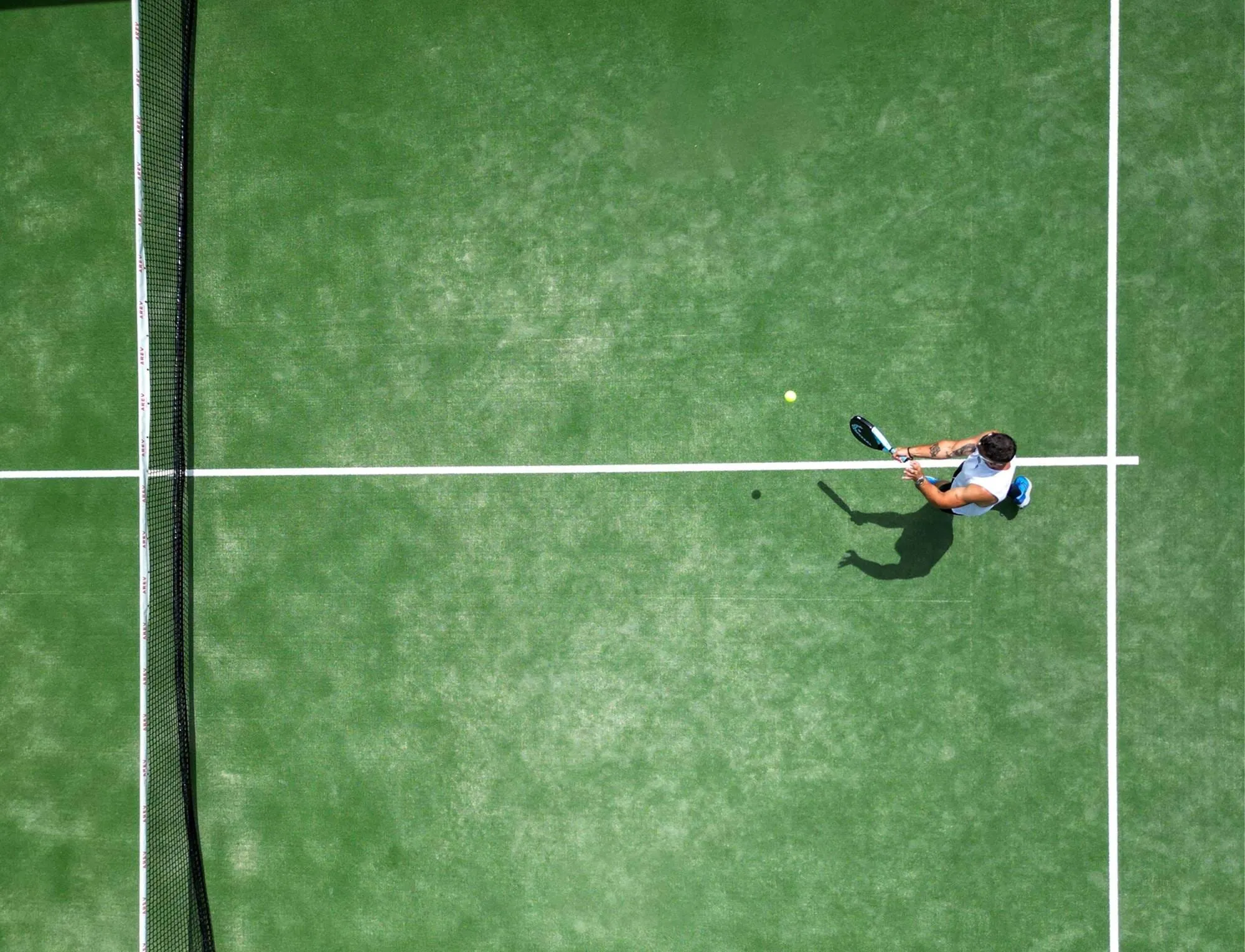 Um jogador a acertar uma bola de ténis num campo de padel verde visto de cima perto da rede.