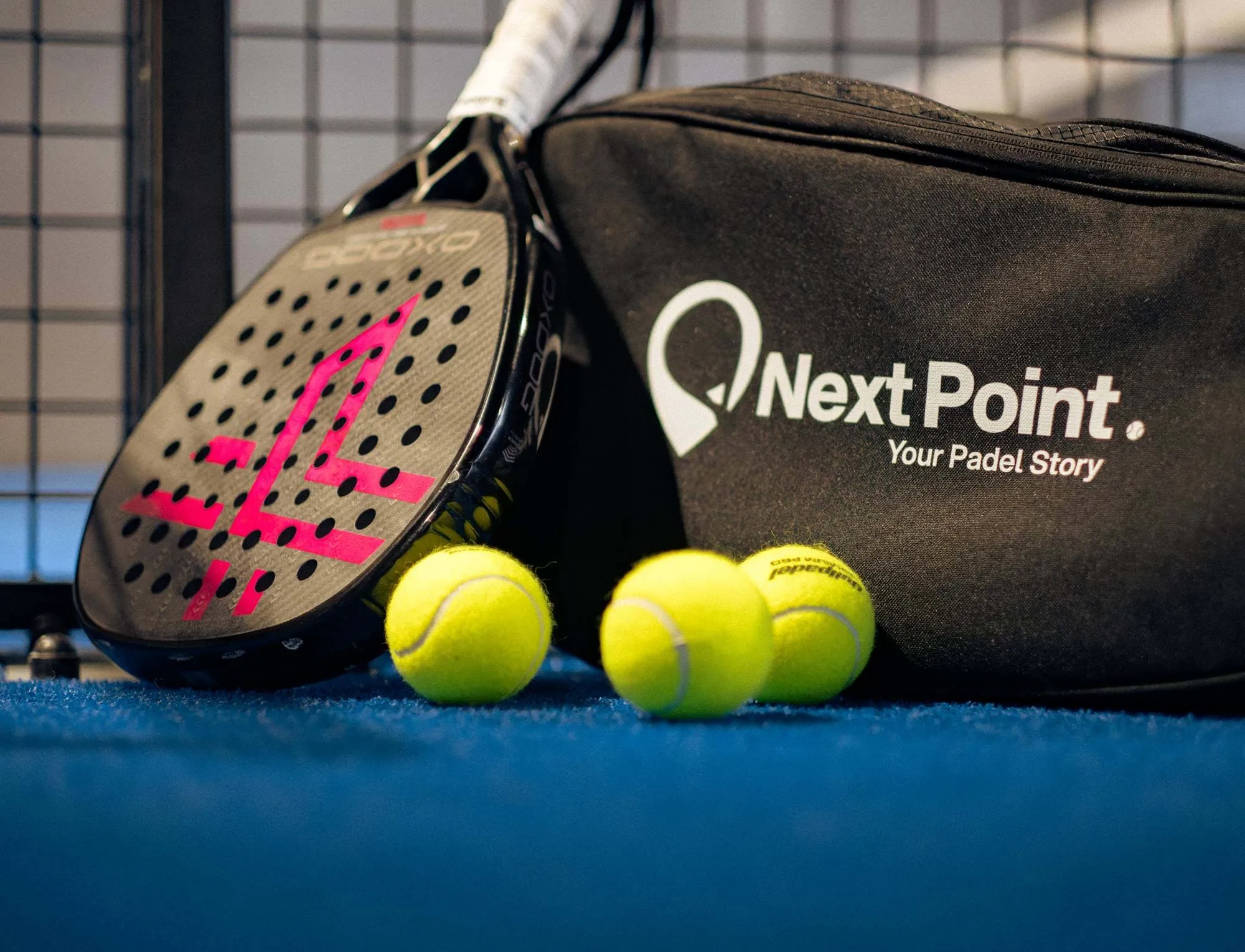 Raquete de padel com logótipo rosa, três bolas de ténis amarelas, e uma bolsa Next Point preta na superfície da quadra azul perto de uma cerca de metal.