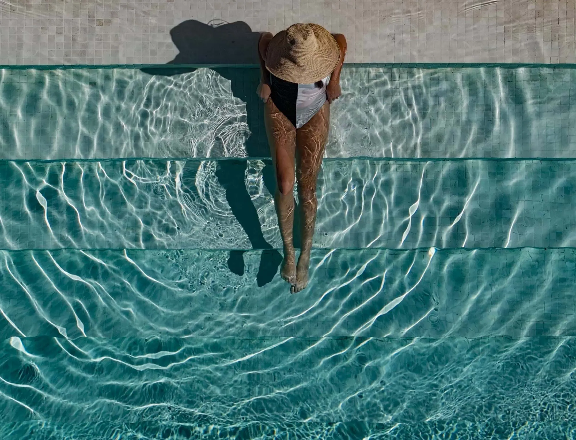Pessoa com chapéu de palha e fato de banho preto e branco sentada nos degraus de azulejos da piscina com as pernas submersas em águas límpidas.
