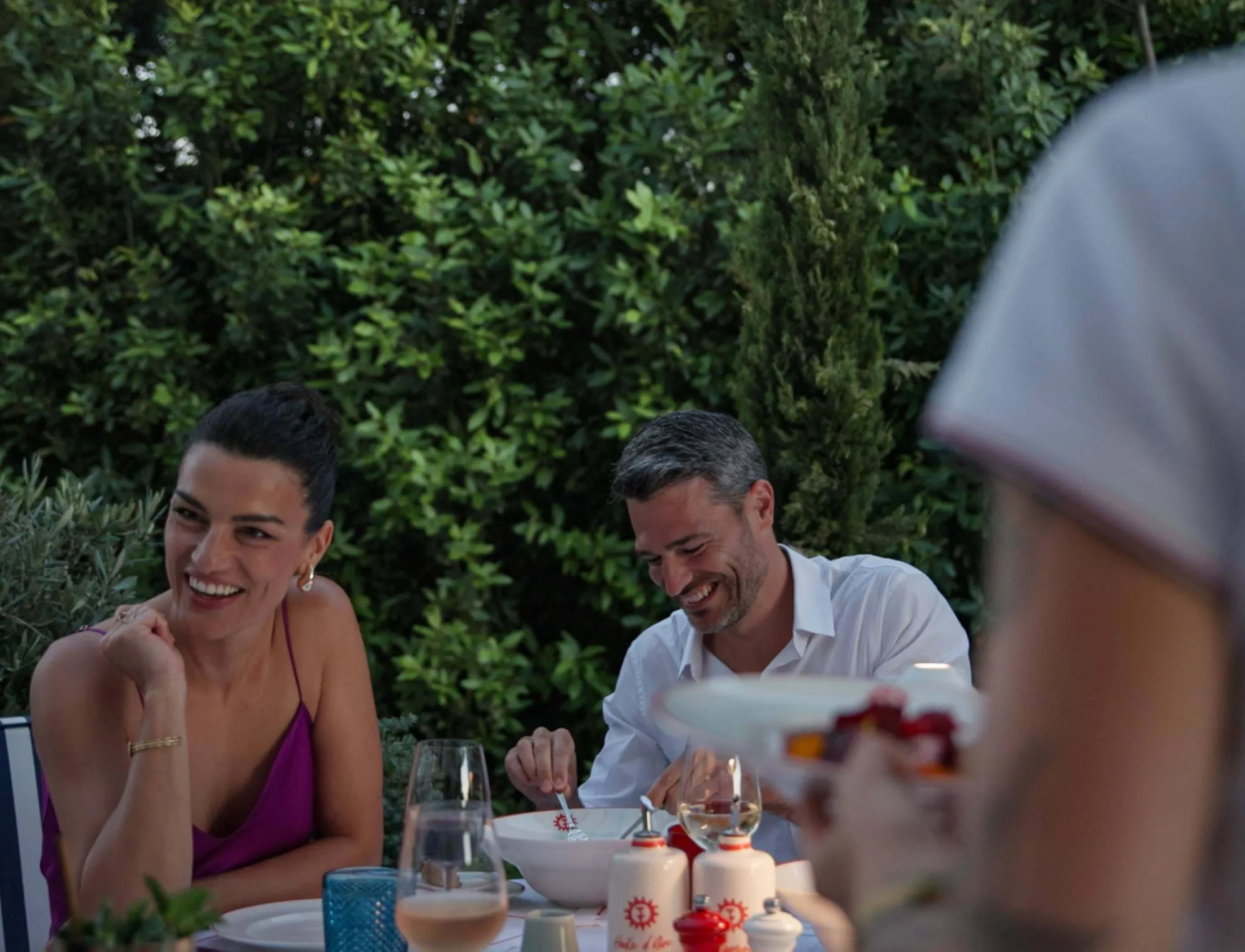 Duas pessoas sorrindo e desfrutando de uma refeição ao ar livre rodeada de vegetação.