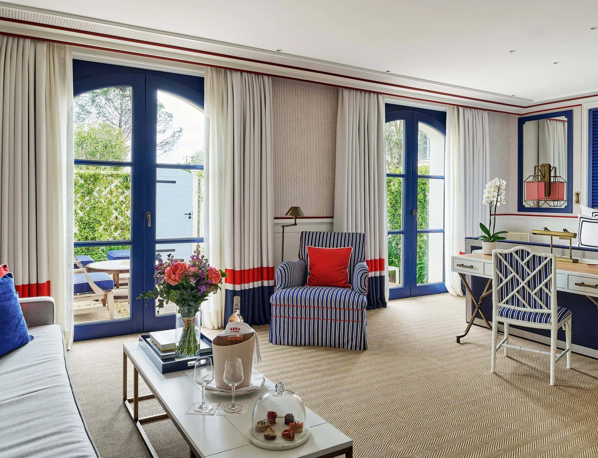 Elegante suite de hotel com decoração azul e branco, poltrona listrada com almofada vermelha, mesa de centro com flores e vinho, e grandes portas de vidro com moldura azul que levam a um terraço.