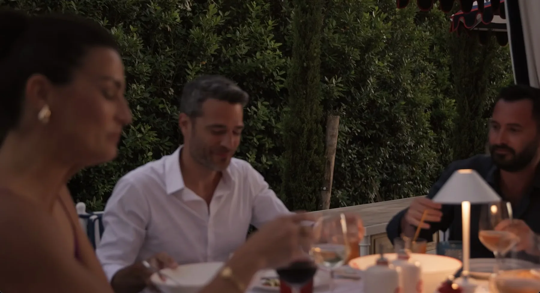 Drei Erwachsene genießen ein Abendessen im Freien an einem Tisch mit Getränken und einer kleinen Lampe, umgeben von grünem Laub.