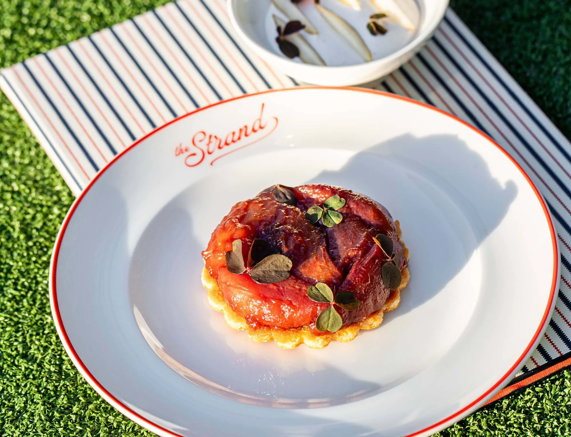 Karamellisierte Fruchttorte, garniert mit grünen Blättern auf einem weißen Teller mit dem Logo „The Strand“, auf gestreiftem Stoff und Gras.