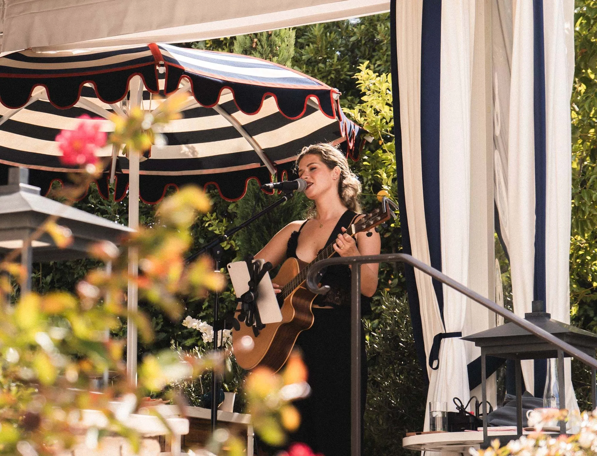Femme chantant et jouant de la guitare acoustique à l'extérieur sous un parapluie rayé noir et blanc avec de la verdure en arrière-plan.