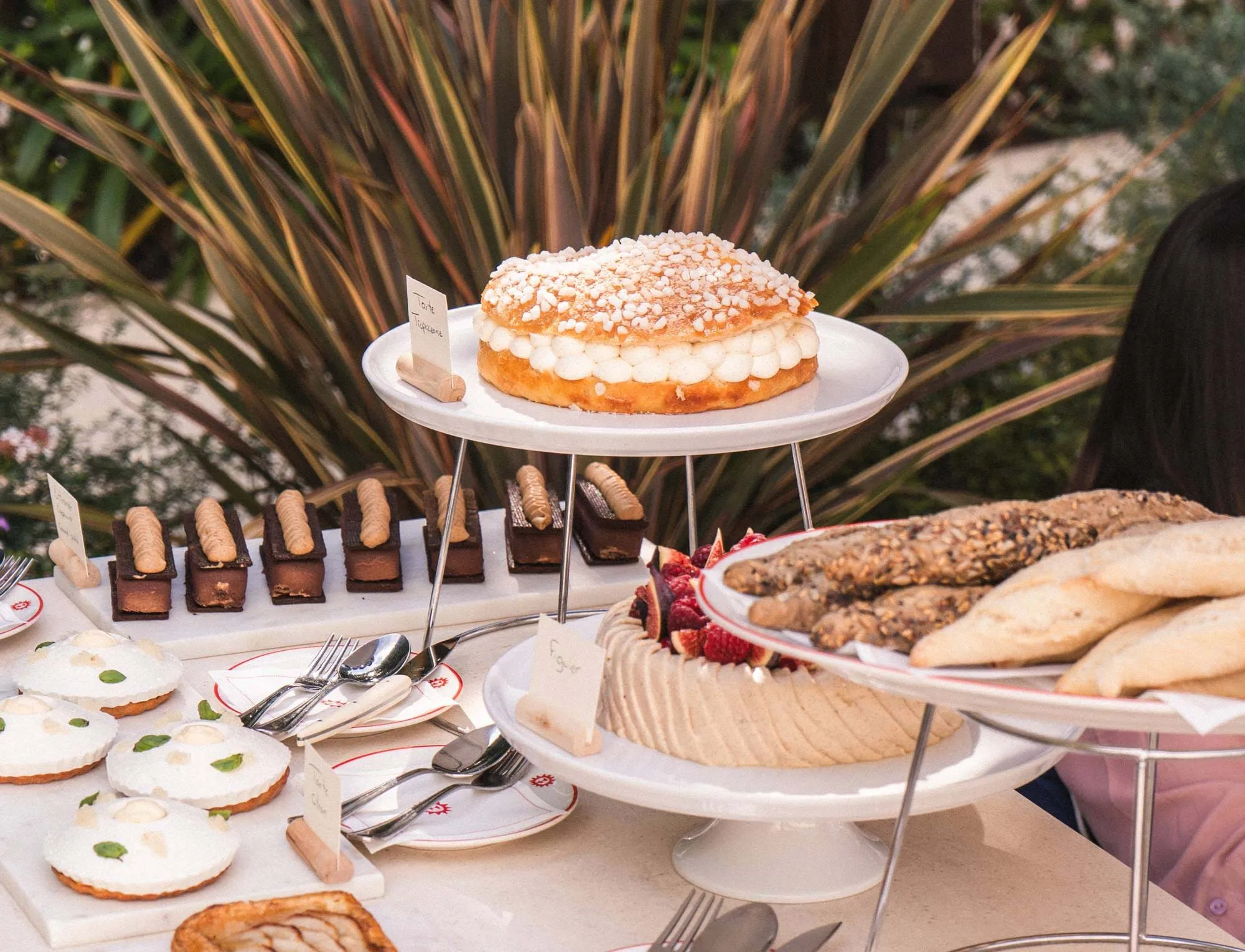 Assortiment de desserts et de pains présentés sur des présentoirs et des assiettes à gâteaux blancs, y compris un gâteau rond à la crème et aux cristaux de sucre, des pâtisseries rectangulaires au chocolat, de petites tartes rondes à la crème blanche et à la menthe, un gâteau garni de baies et des baguettes aux graines.