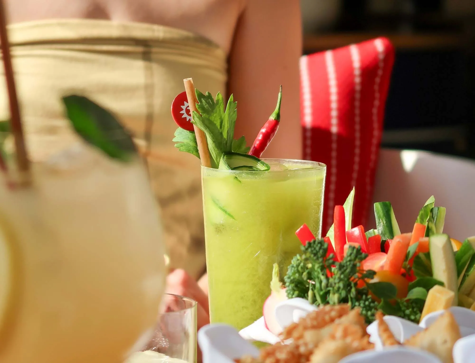 Cocktail verde guarnecido com aipo, pepino e pimenta vermelha ao lado de um prato de palitos de legumes frescos e lanches.