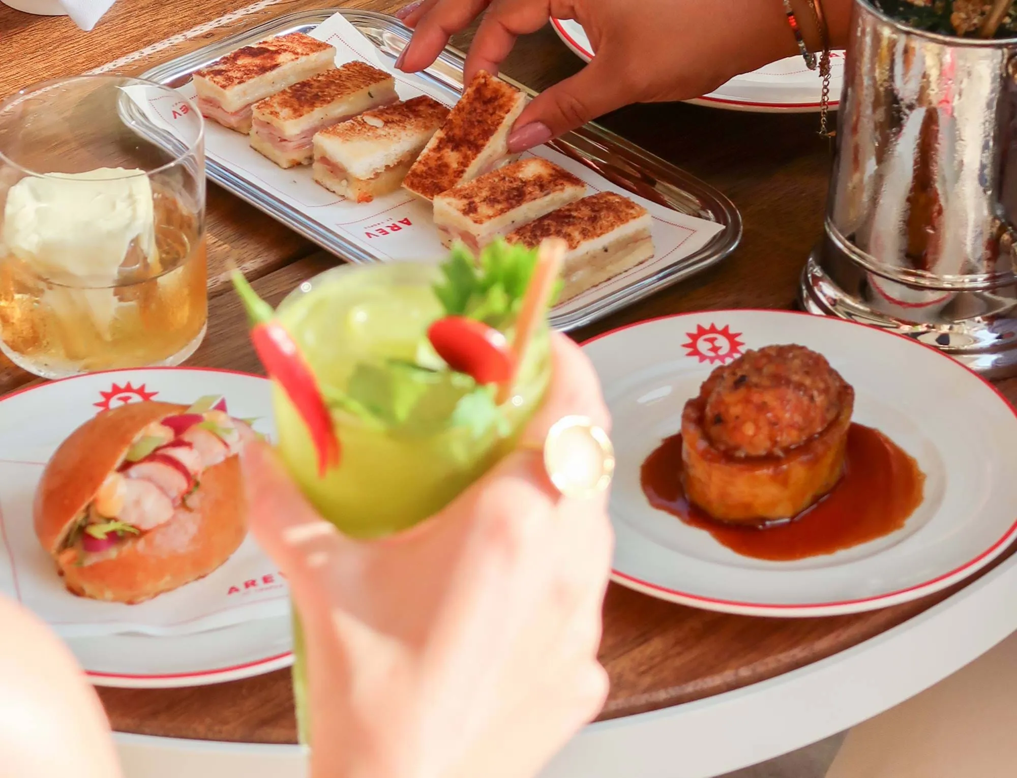 Uma mão segurando um cocktail verde guarnecido com pimentas vermelhas e ervas, com pratos de aperitivos gourmet e um copo de whisky sobre uma mesa de madeira.