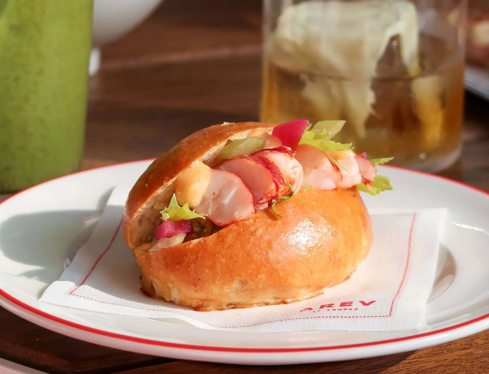 Rolo de lagosta com alface e molho num coque vitrificado num prato branco com um guardanapo, com bebidas ao fundo.