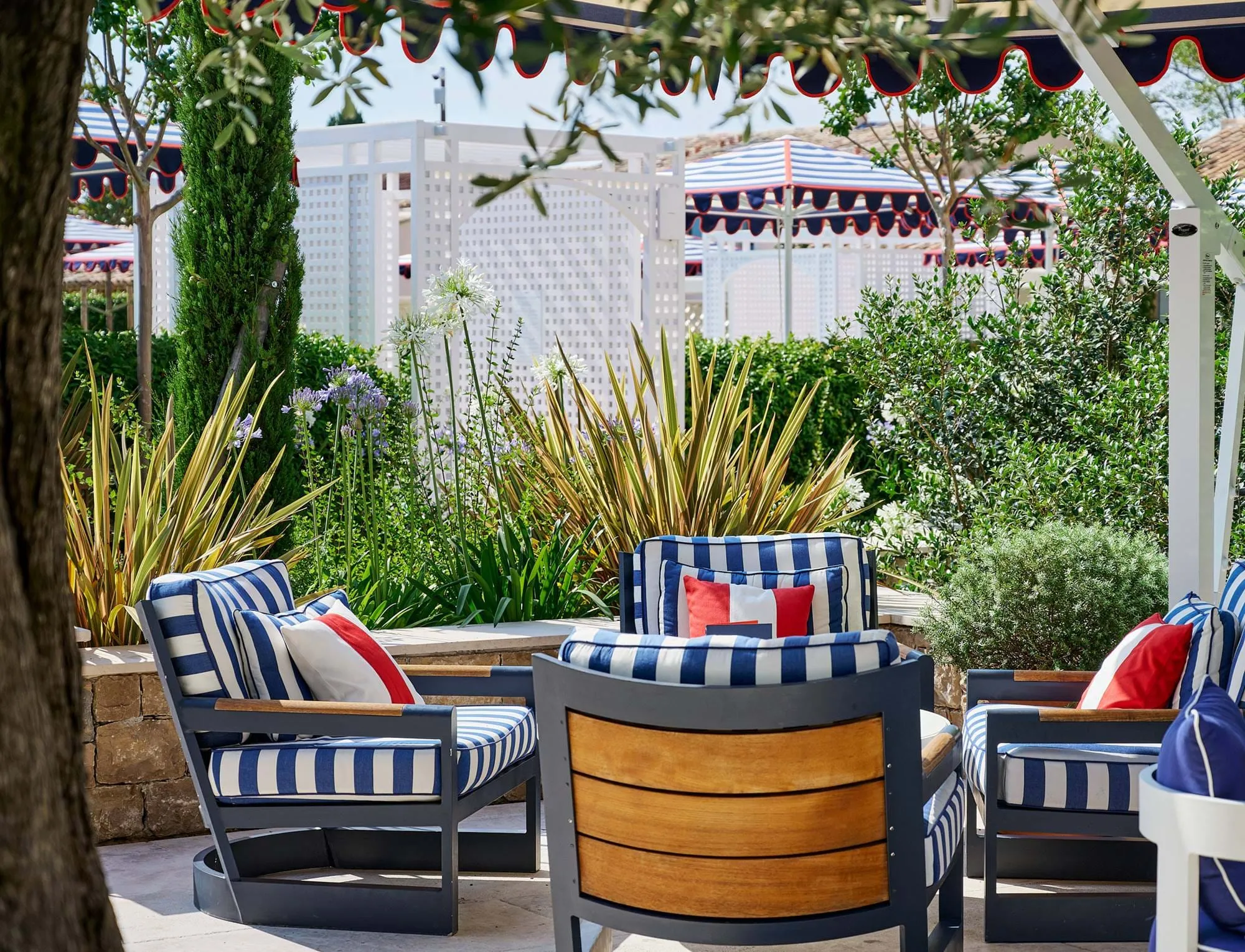 Área de estar exterior com almofadas listradas azul-marinho e branco e almofadas de acento vermelho rodeadas por plantas verdes e estruturas de jardim brancas sob toldos listrados.