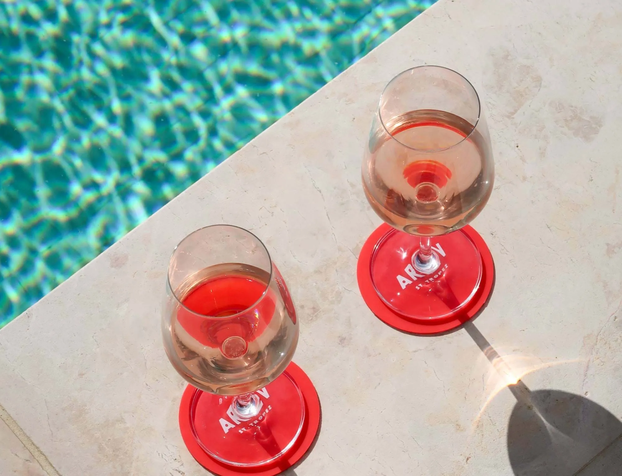 Dois copos de vinho rosé nas montanhas-russas vermelhas ao lado de uma piscina azul sobre uma superfície de pedra iluminada pelo sol.