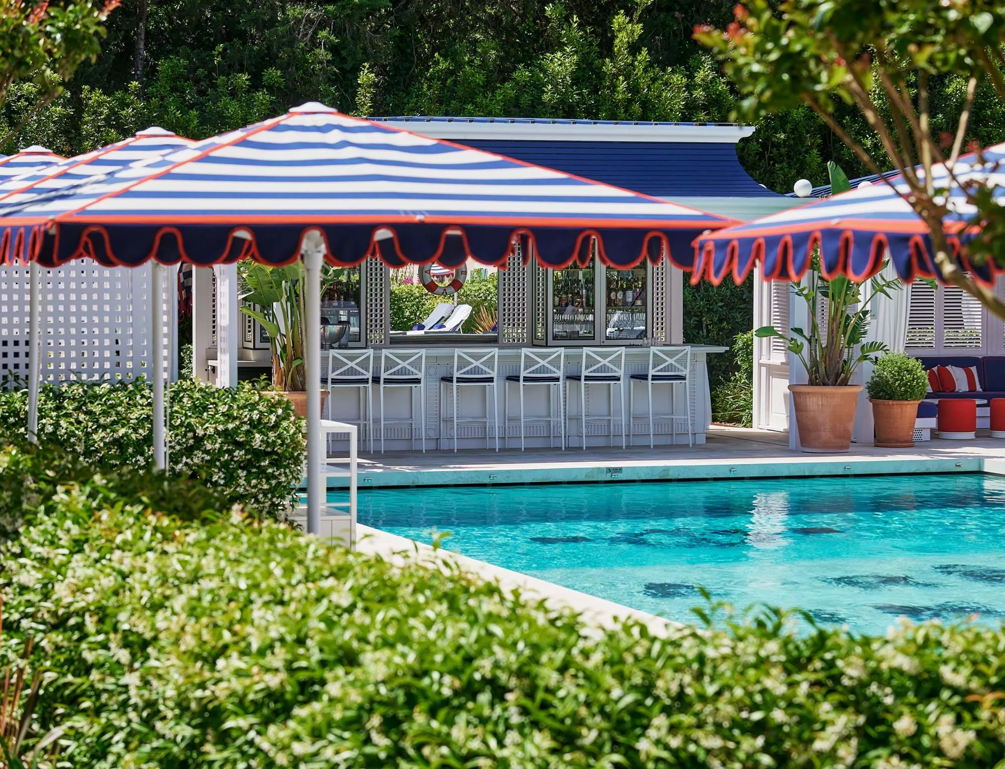 Bar junto à piscina com bancos brancos e guarda-chuvas listrados em azul e branco rodeado de vegetação.