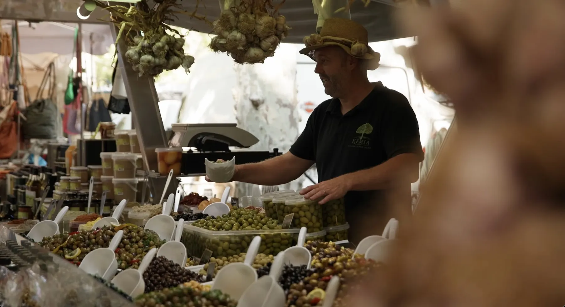 Verkäufer mit Strohhut, der Oliven an einem Marktstand im Freien mit verschiedenen Oliven und Gläsern serviert.