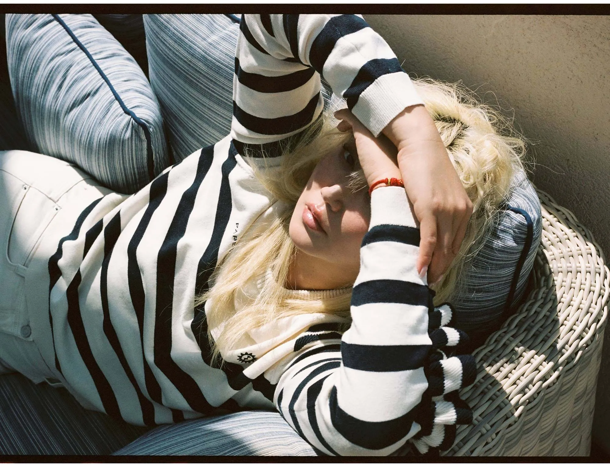 Mulher loira reclinada numa cadeira de vime com almofadas azuis listradas, vestindo um suéter listrado preto e branco e a proteger os olhos com o braço.