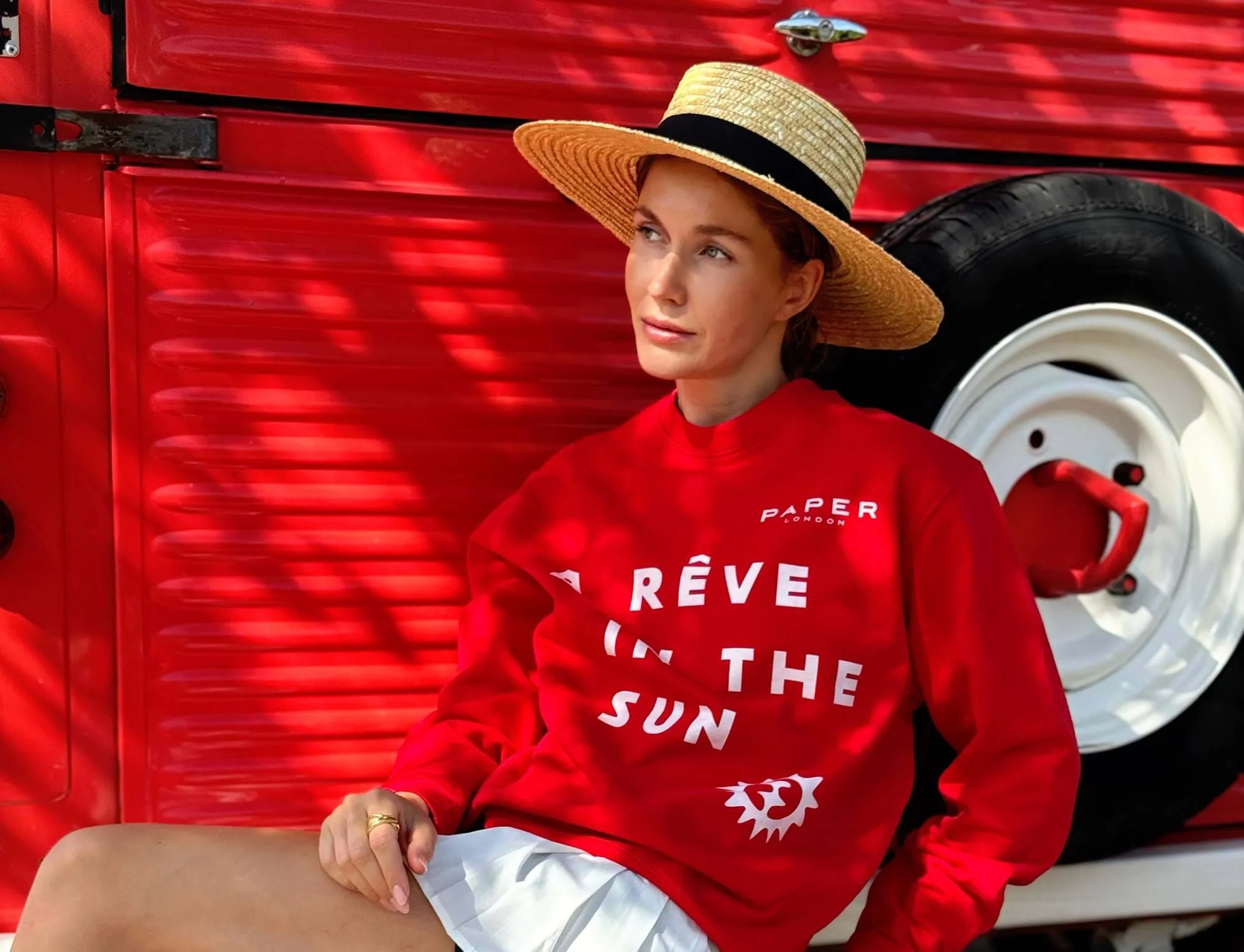 Woman wearing a straw hat and red sweatshirt with text sitting against a red vehicle with a large white and red wheel.