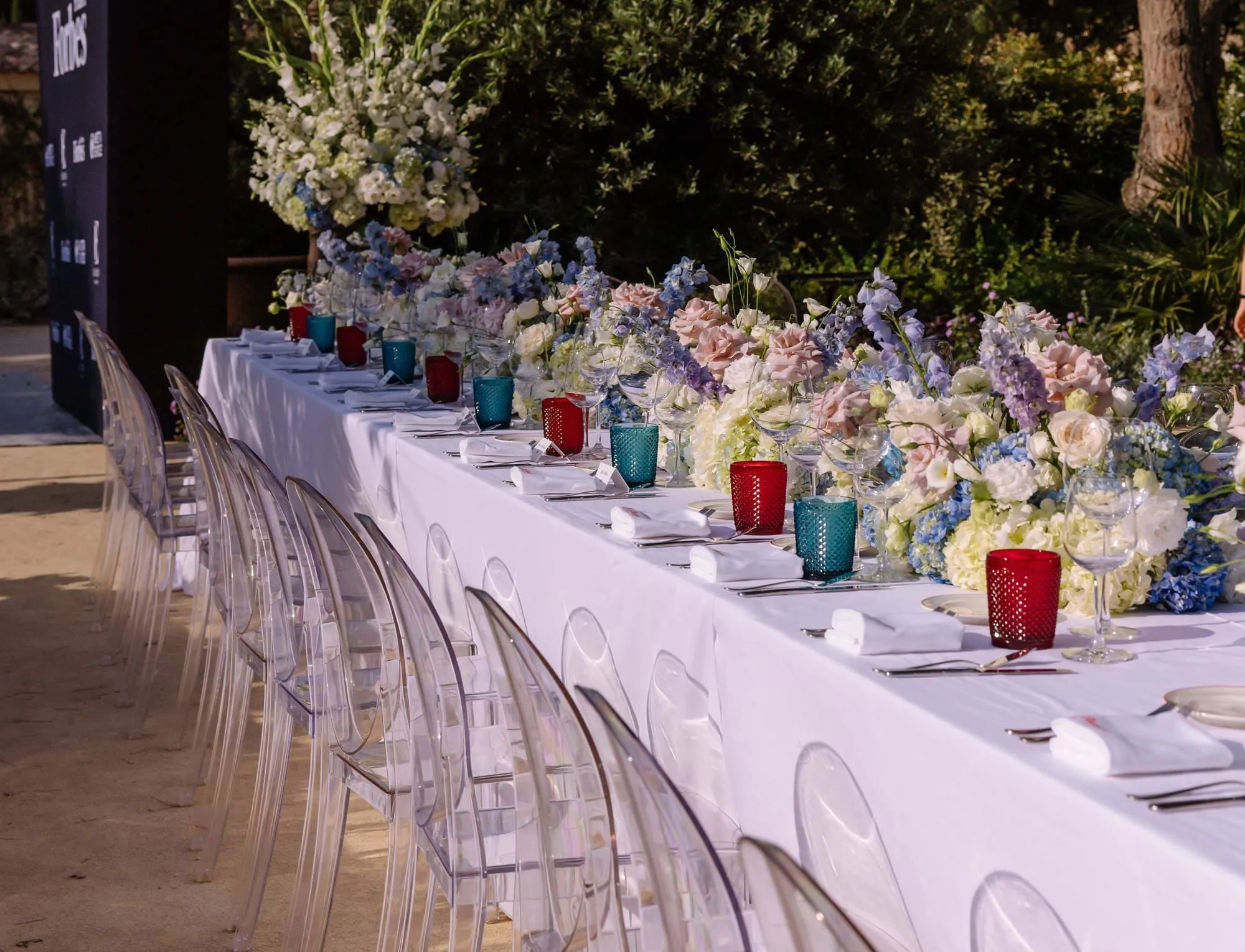 Mesa de jantar longa ao ar livre com toalha de mesa branca, cadeiras transparentes, peças centrais florais pastel e castiçais de vidro vermelho e azul.