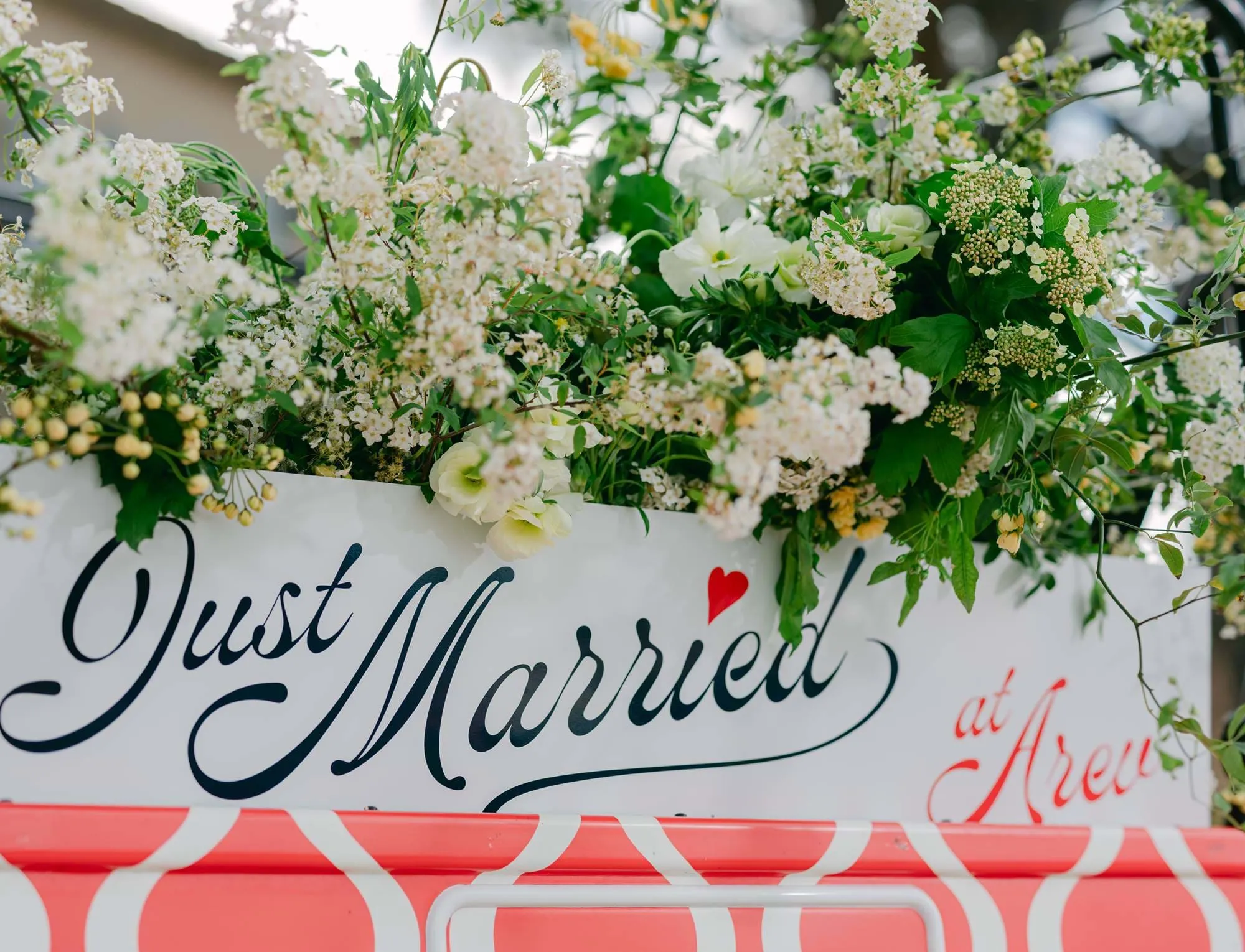 Sinal branco com caligrafia preta e vermelha onde se lê 'Just Married at Arev' adornado com exuberantes arranjos florais brancos e verdes.