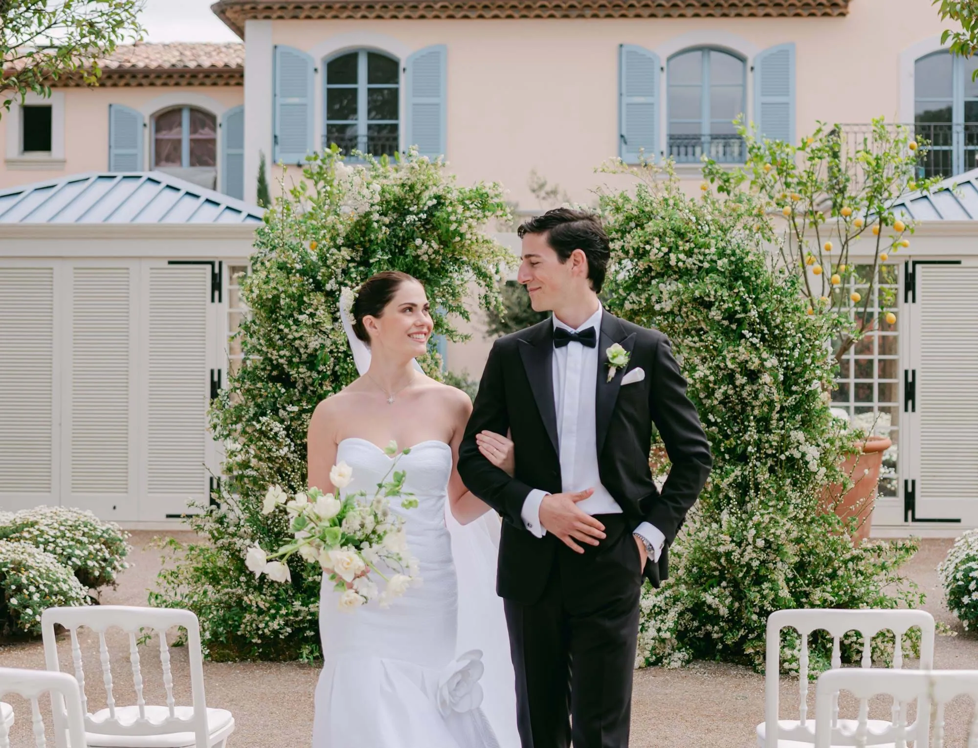Noiva em vestido branco sem alças segurando buquê de braços dados de braço a braço com o noivo em smoking preto, em pé ao ar livre com vegetação e um edifício com persianas azuis atrás.