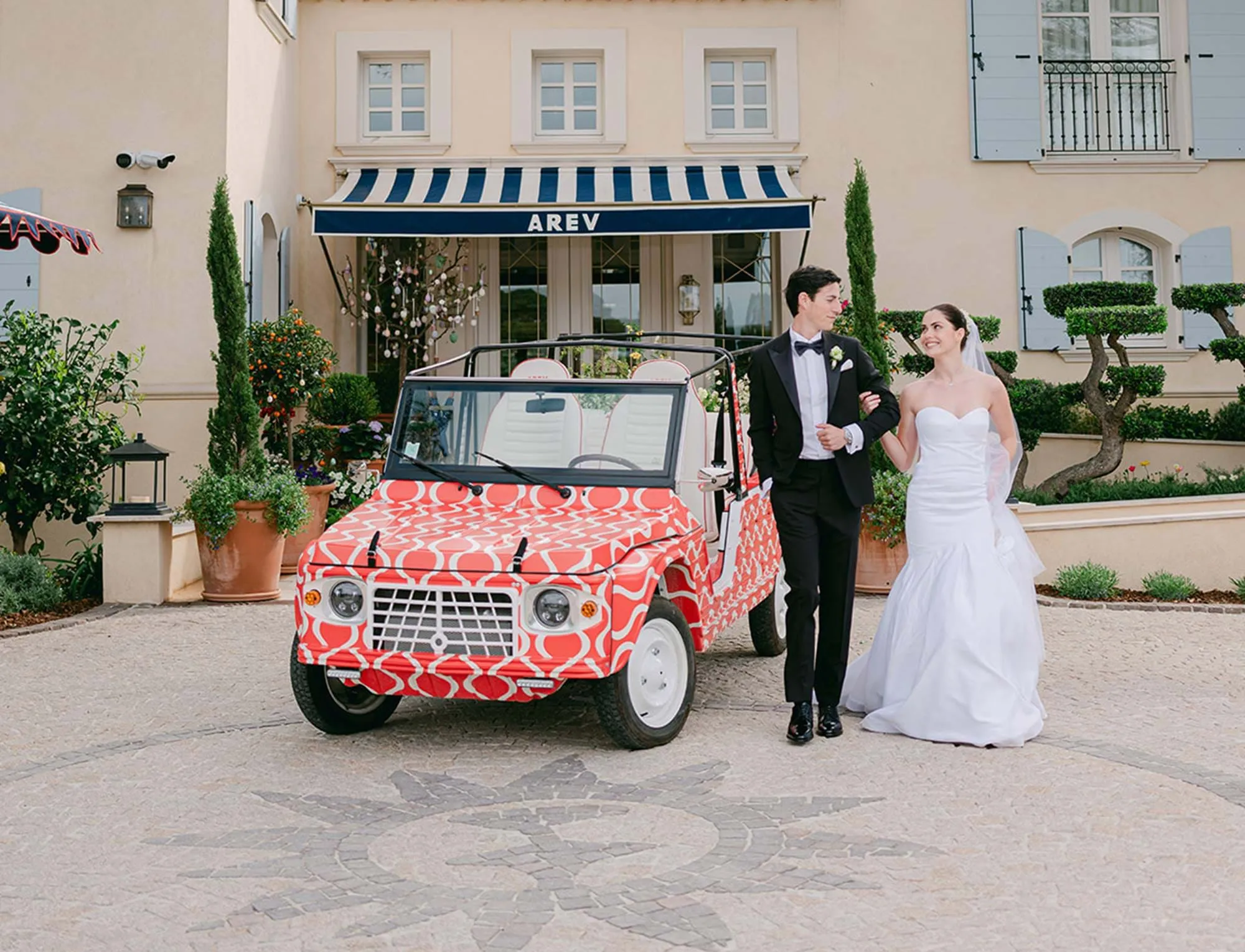 Noiva e noivo sorrindo e segurando os braços enquanto estão ao lado de um carro conversível vintage com estampar vermelho em frente a um edifício com um toldo listrado azul e branco.