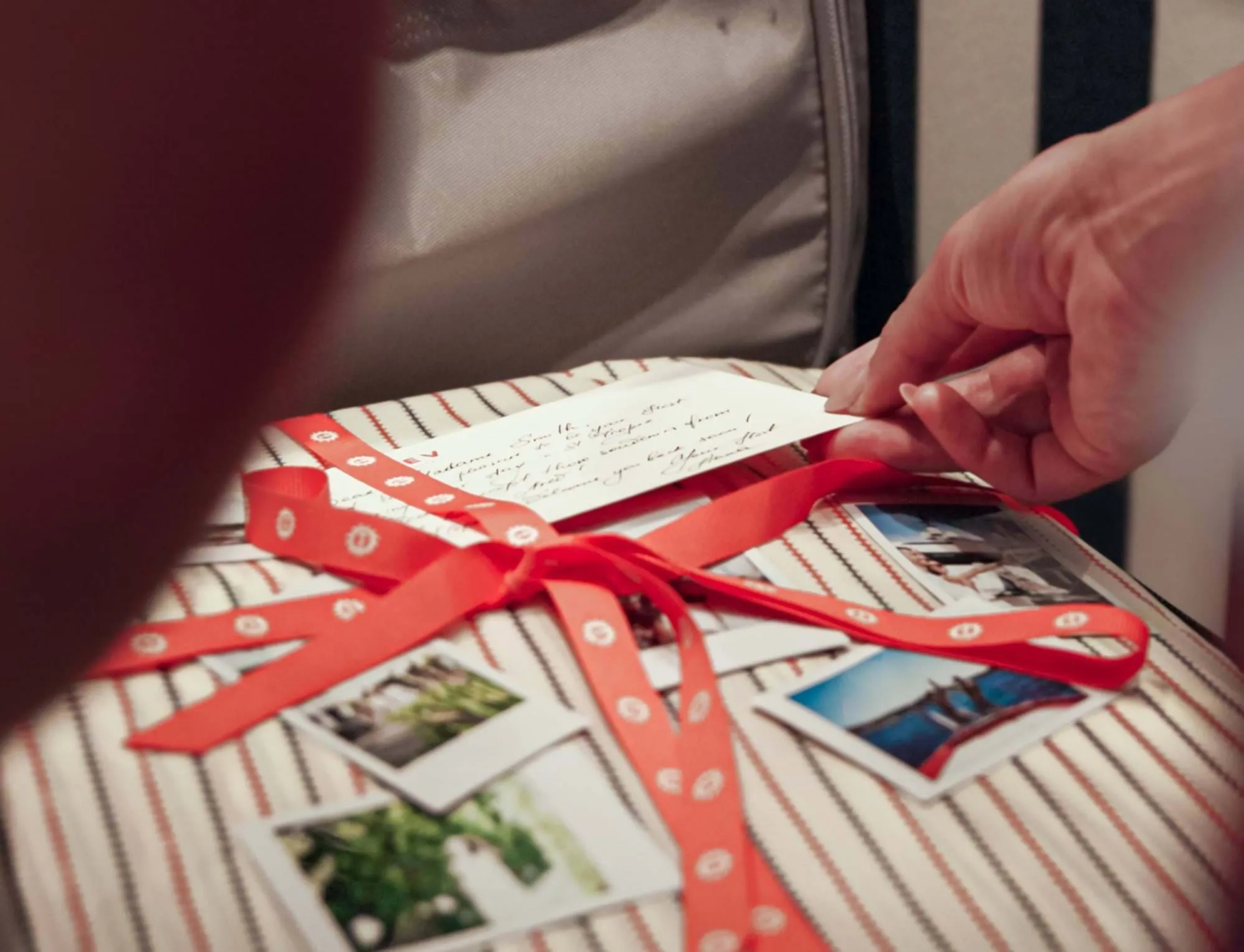Mão segurando uma nota manuscrita colocada sobre uma superfície de tecido listrado com fita vermelha e várias fotos impressas.