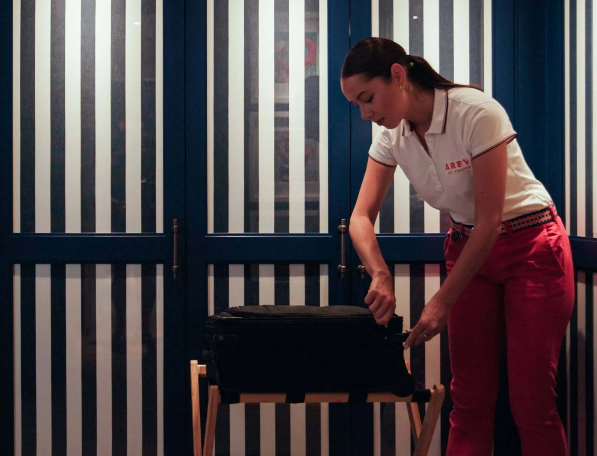 Uma mulher de polo branco e calças vermelhas a abrir uma mala preta num bagageiro de madeira em frente a marinho e portas listradas brancas.