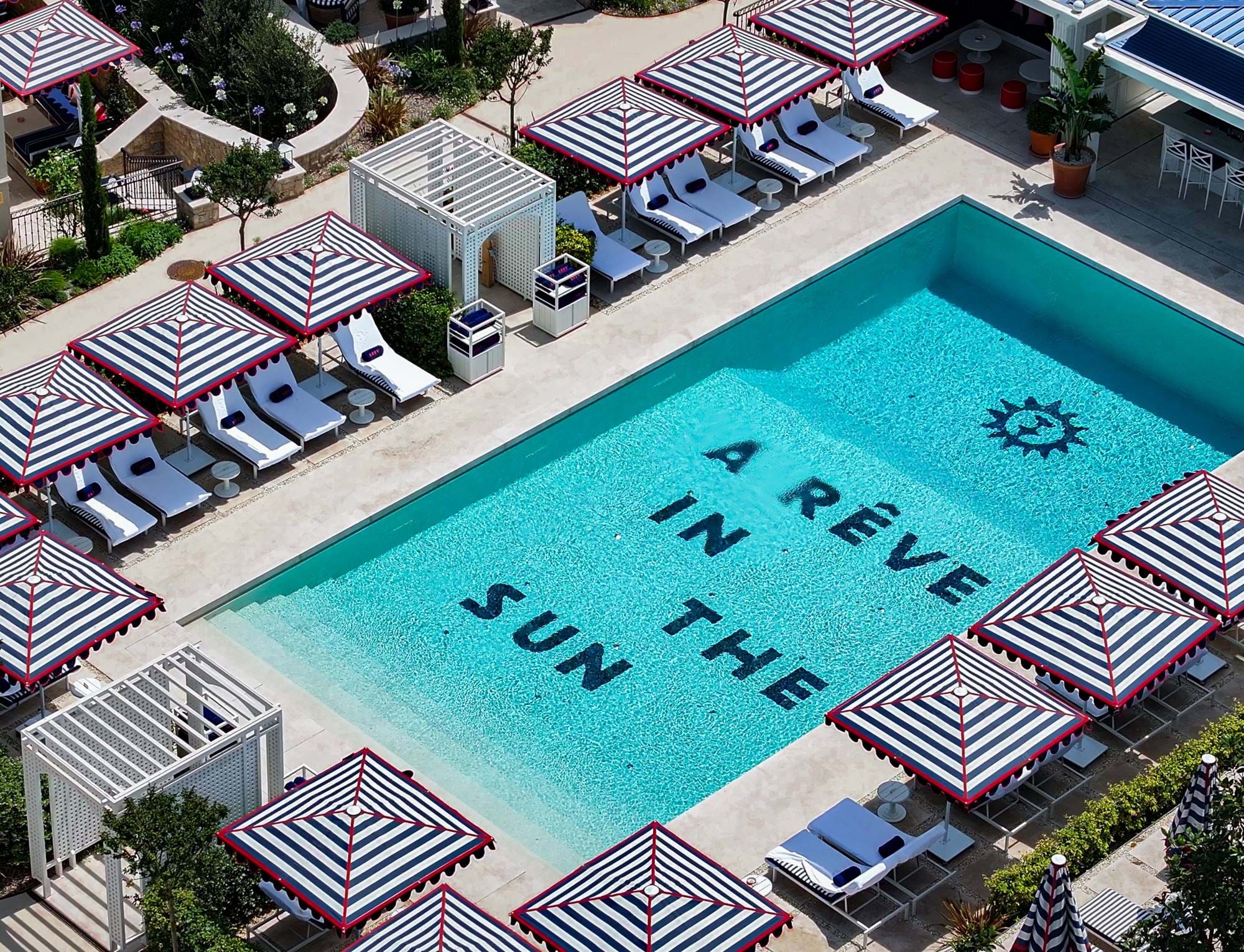 Piscina exterior com água azul rodeada de espreguiçadeiras brancas e guarda-sóis com riscas marinhas, com as palavras 'A RÊVE IN THE SUN' visíveis debaixo d'água.