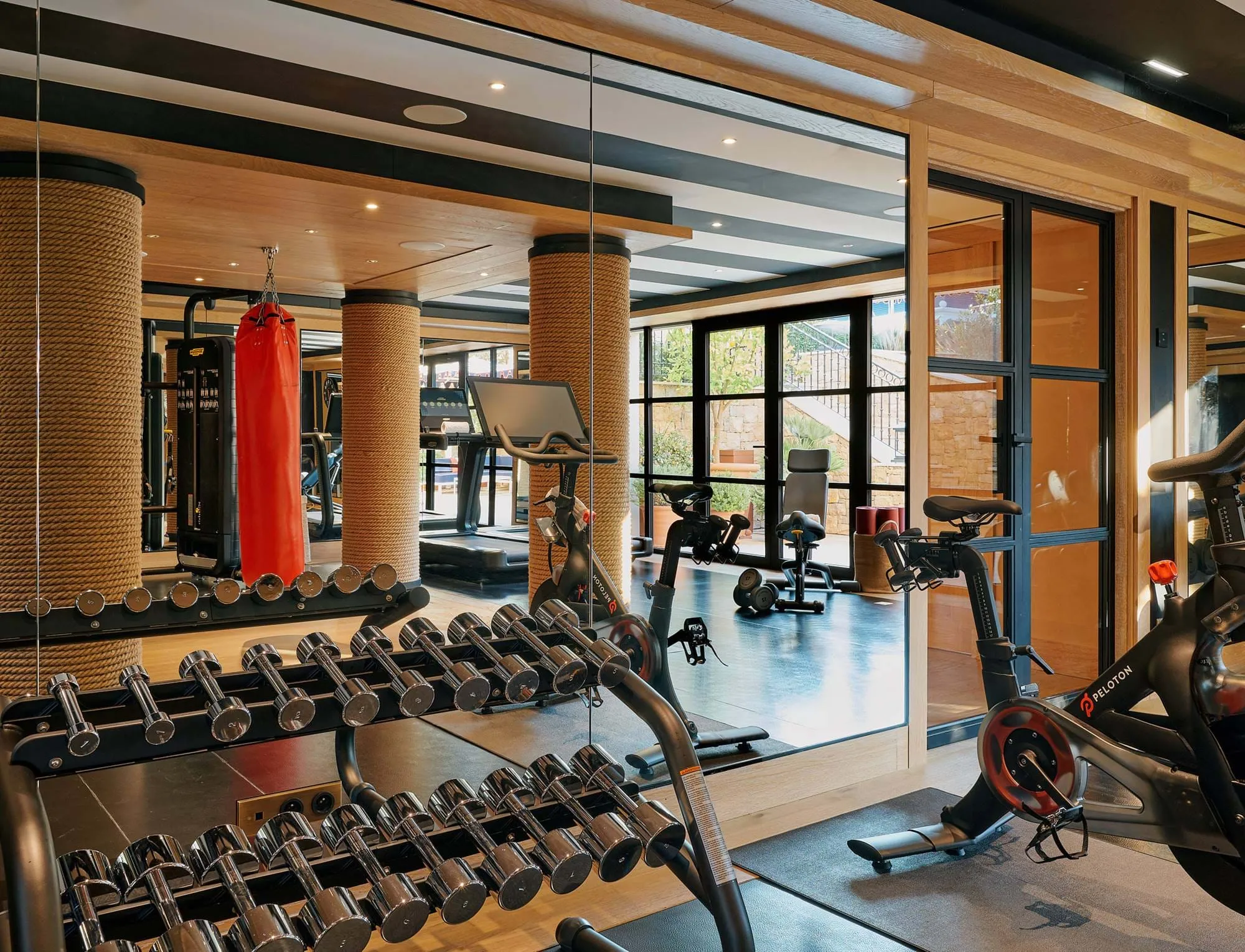 Sala de ginásio moderna com suporte para halteres, bicicleta Peloton, saco de pancadas e grandes espelhos que refletem o equipamento de treino.