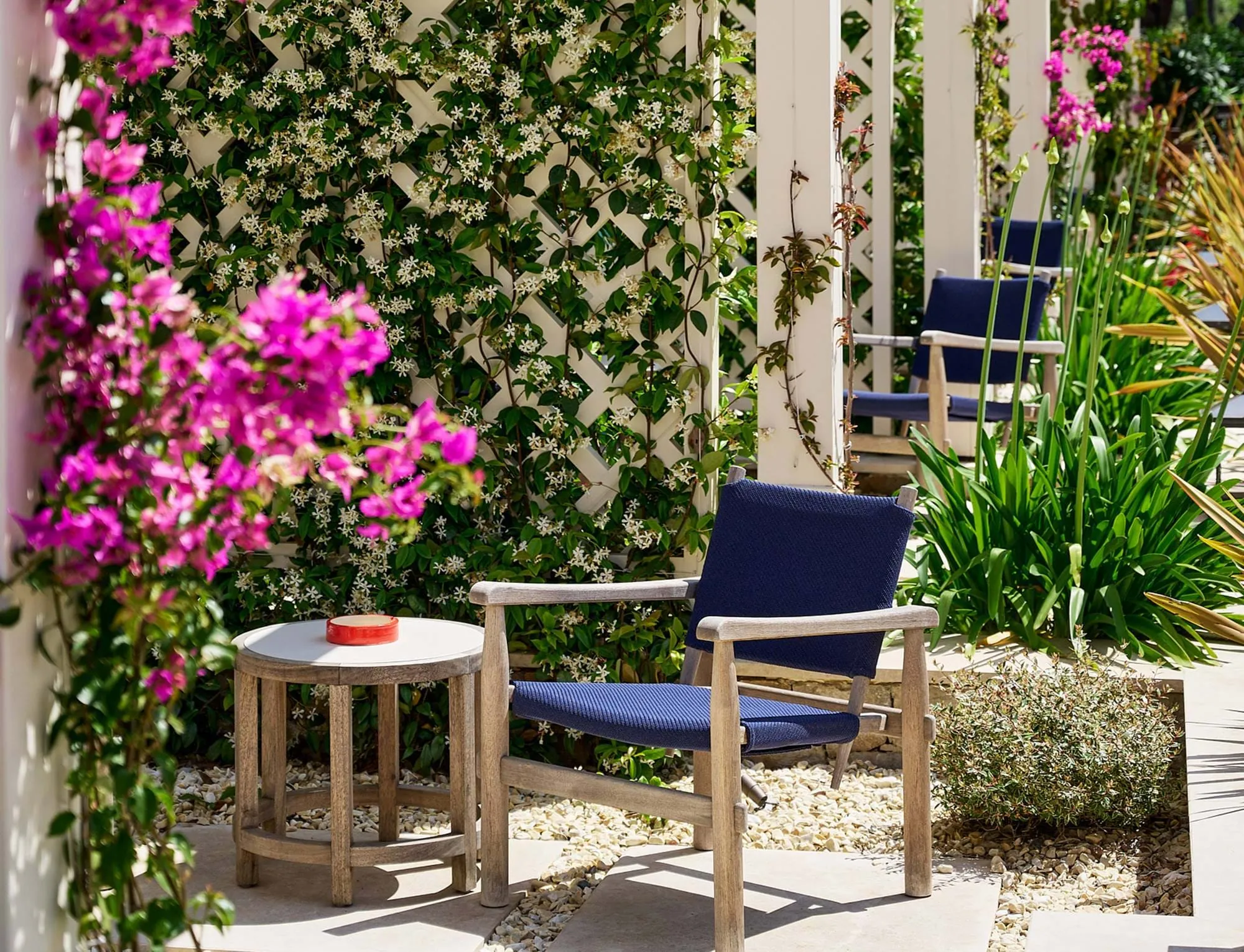 Área de estar exterior com cadeiras de madeira com almofadas azuis, uma pequena mesa redonda e plantas com flores num pátio.