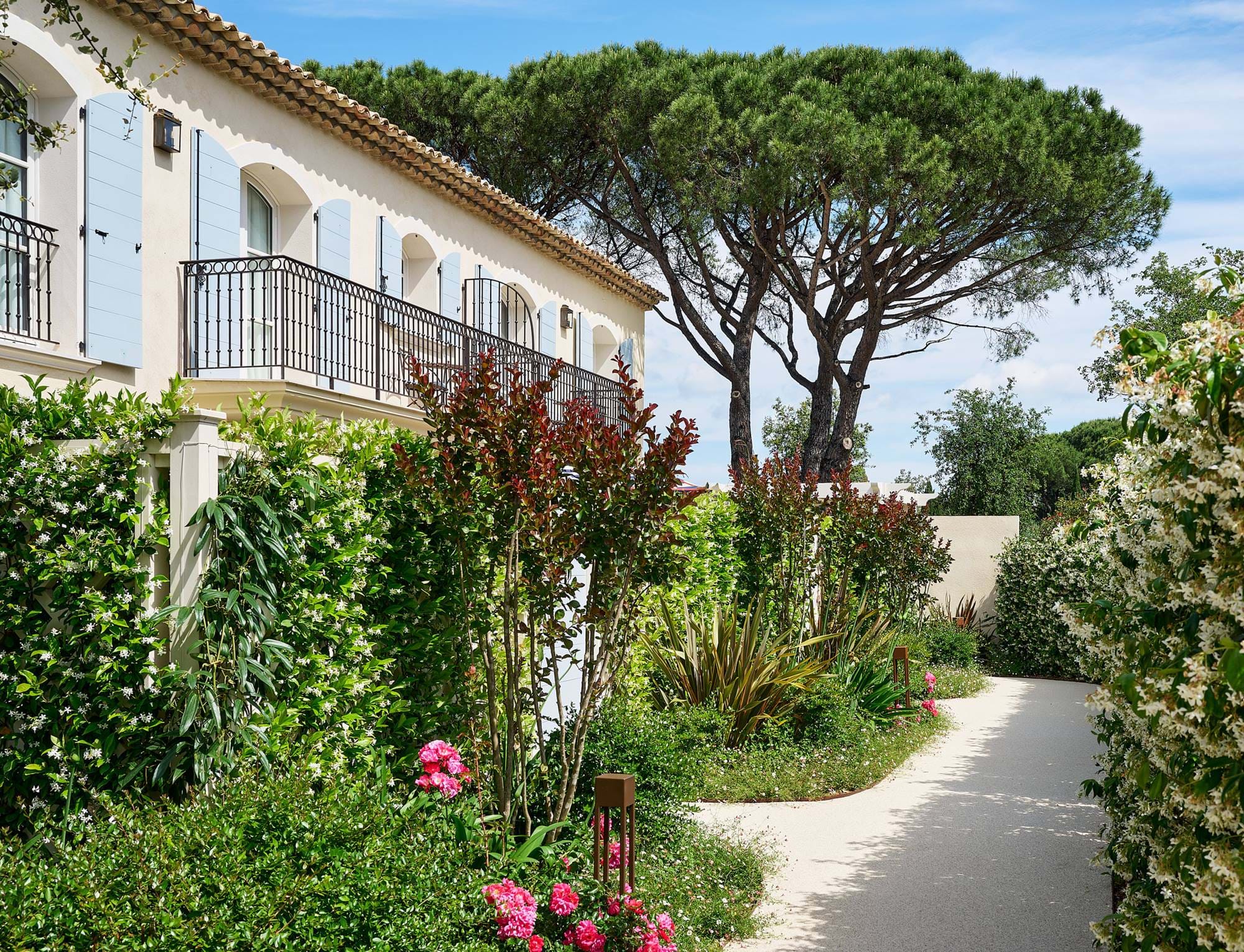 Caminho de jardim iluminado pelo sol, forrado por arbustos verdes e flores rosa ao lado de um edifício de cor creme com portadas azuis e varandas pretas.