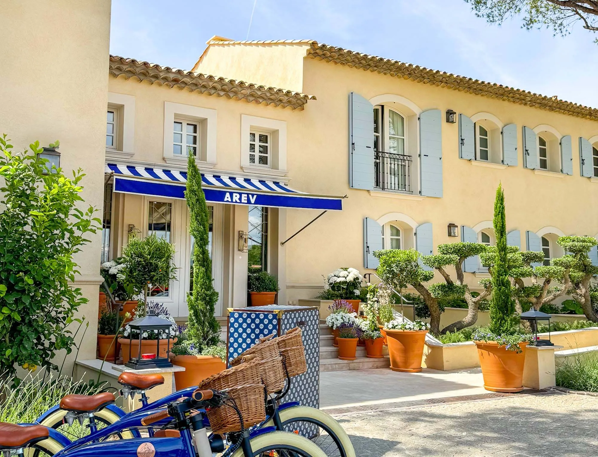 Des vélos bleus avec des paniers tressés garés à l'extérieur d'un bâtiment beige avec des volets bleus et un auvent rayé bleu étiqueté AREV.