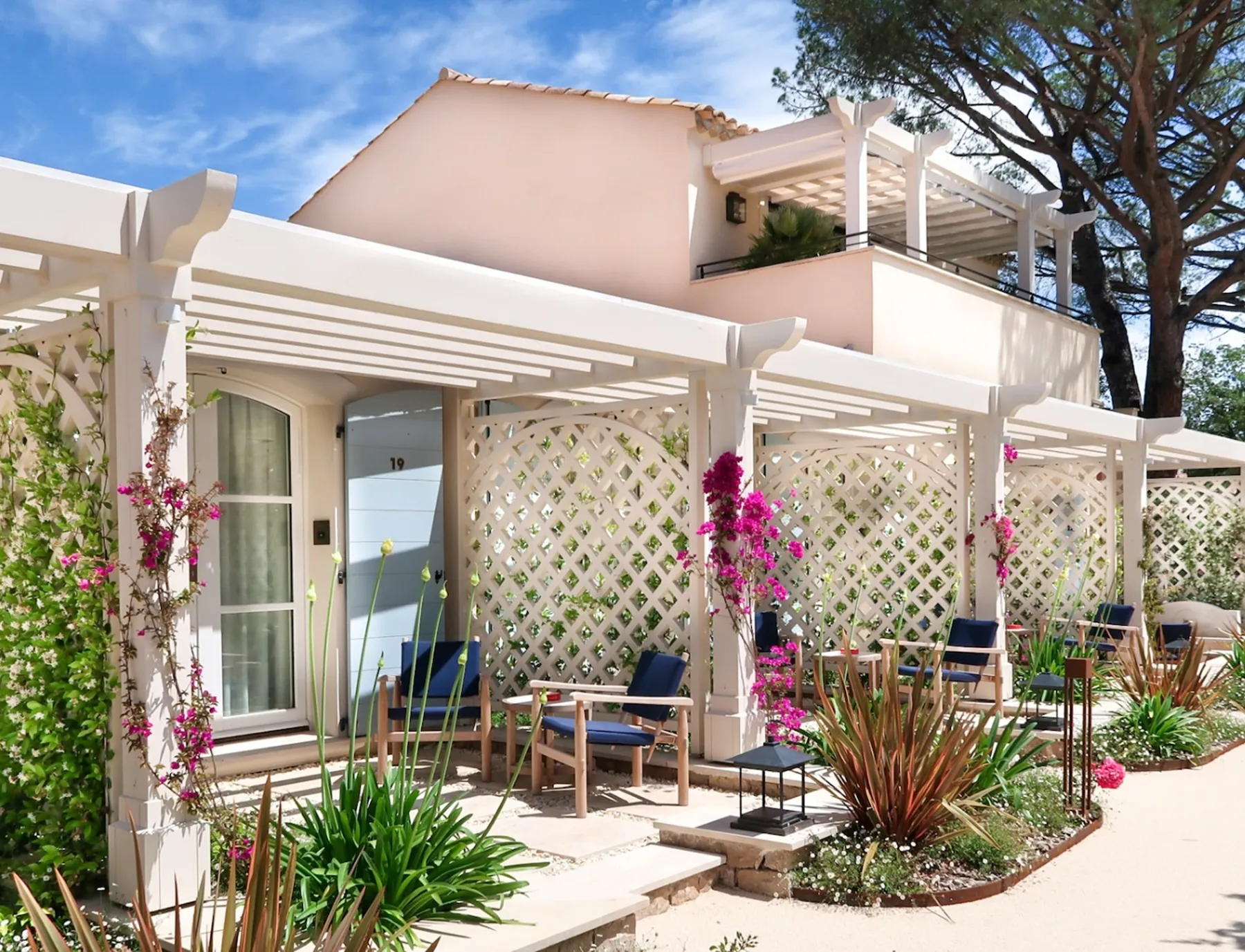 Área de pátio exterior ensolarada com pérgulas brancas, cadeiras de madeira com almofadas azuis e plantas floridas em frente a um edifício de cor clara.