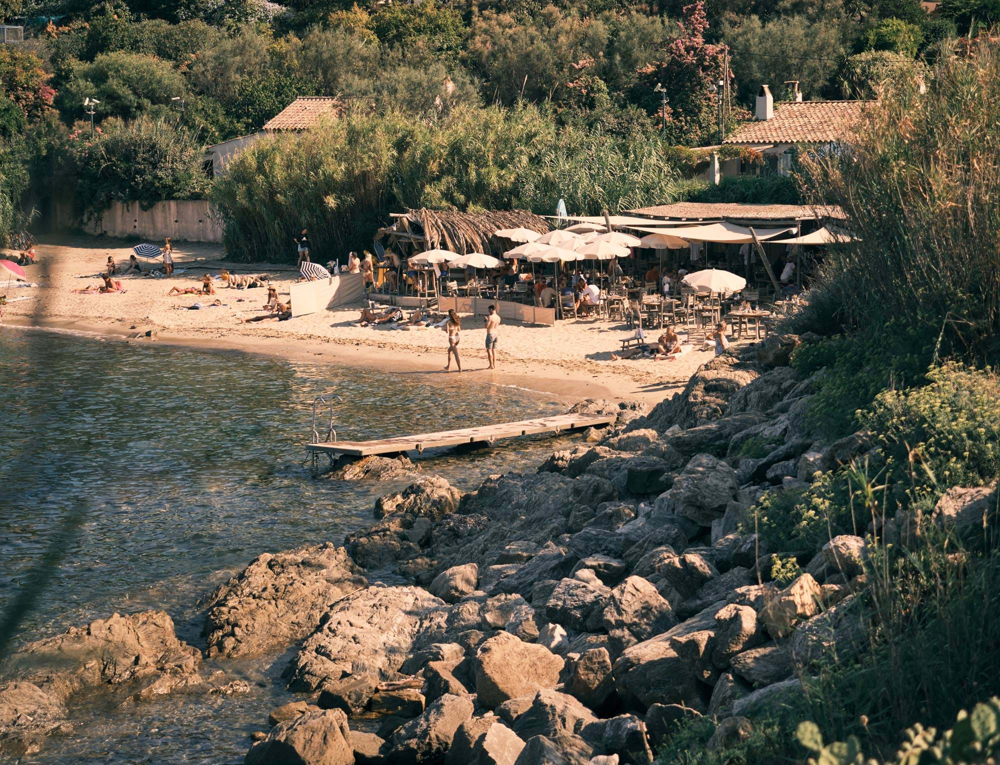 Praia de areia com pessoas a apanhar sol e um café à beira-mar com guarda-sóis e cadeiras rodeado de vegetação e costa rochosa.