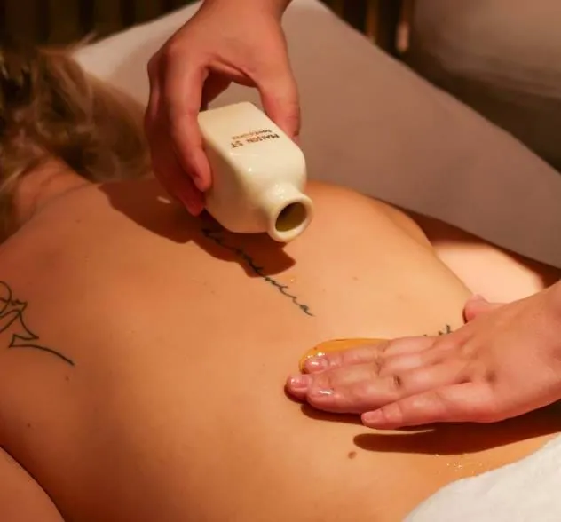 Pessoa a receber uma massagem nas costas com óleo a ser derramado de uma garrafa e espalhado à mão, mostrando tatuagens nas costas.