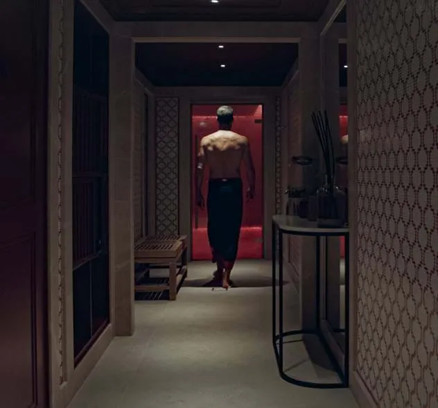 Homem sem camisas andando por um corredor mal iluminado com paredes estampadas e um banco à esquerda.