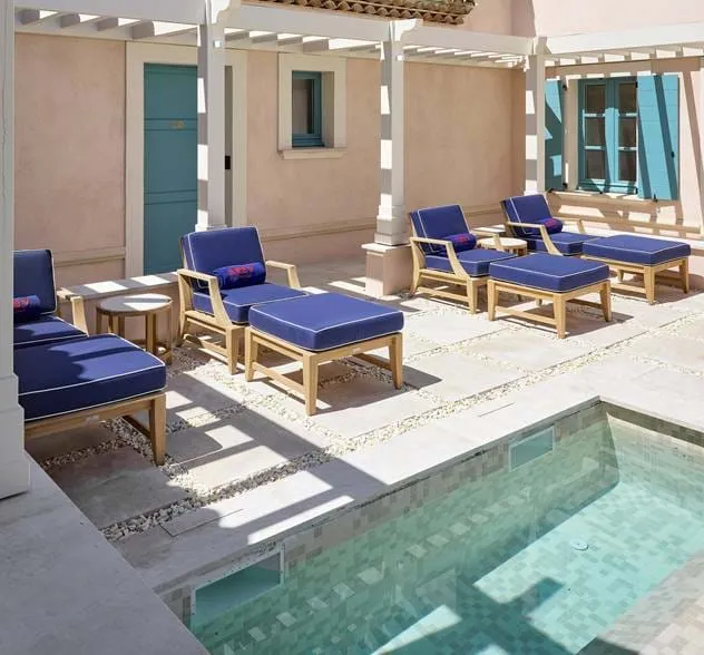 Área exterior junto à piscina com espreguiçadeiras de madeira com almofadas azuis e pequenas mesas laterais sob uma pérgola branca.