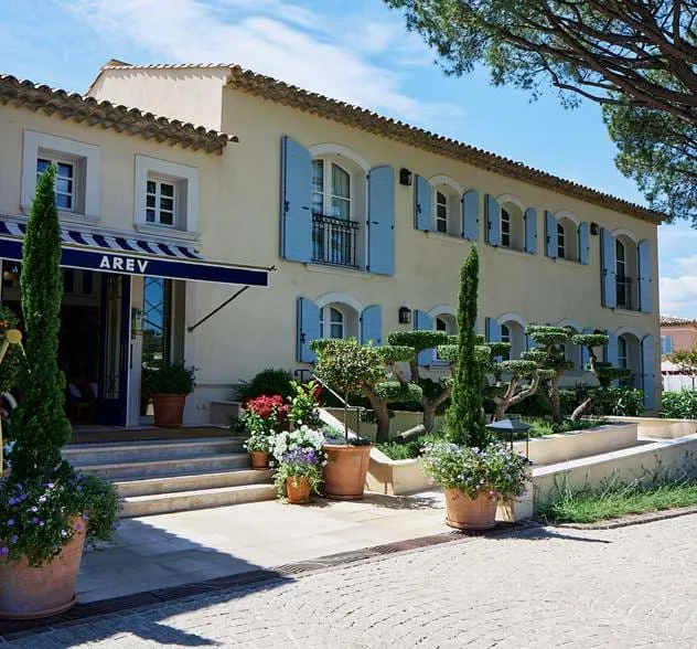 Edifício de estilo mediterrâneo com paredes amarelas pálidas, persianas azuis e vasos de plantas na entrada sob um toldo rotulado AREV.