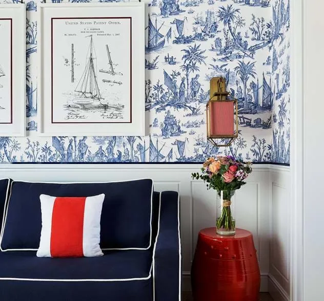 Sofá azul marinho com tubulação branca e uma almofada vermelha, branca e azul ao lado de uma mesa lateral vermelha com um vaso de flores, encostado em papel de parede náutico branco e azul com desenhos de patentes emoldurados de veleiros.